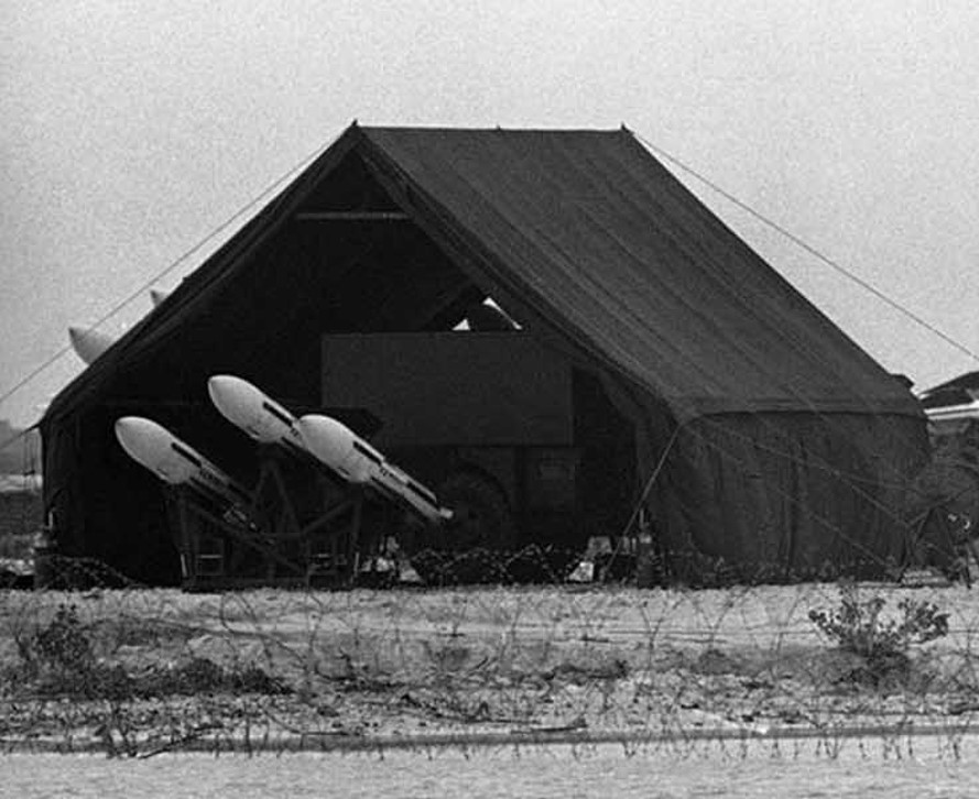 Three white missiles on launchers beneath a large military tent behind barbed wire