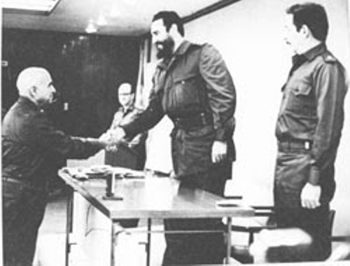 Black-and-white photograph of a handshake across a table between a man in a dark jacket and a bearded officer in fatigues