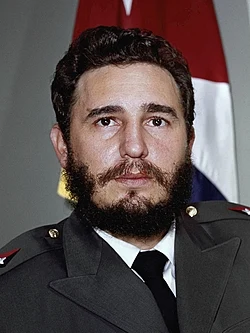 Colour portrait of Fidel Castro in an olive military uniform with a Cuban flag behind him