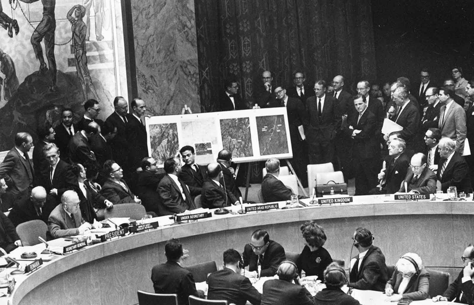 Black-and-white photograph of the United Nations Security Council chamber with aerial reconnaissance photographs displayed on an easel