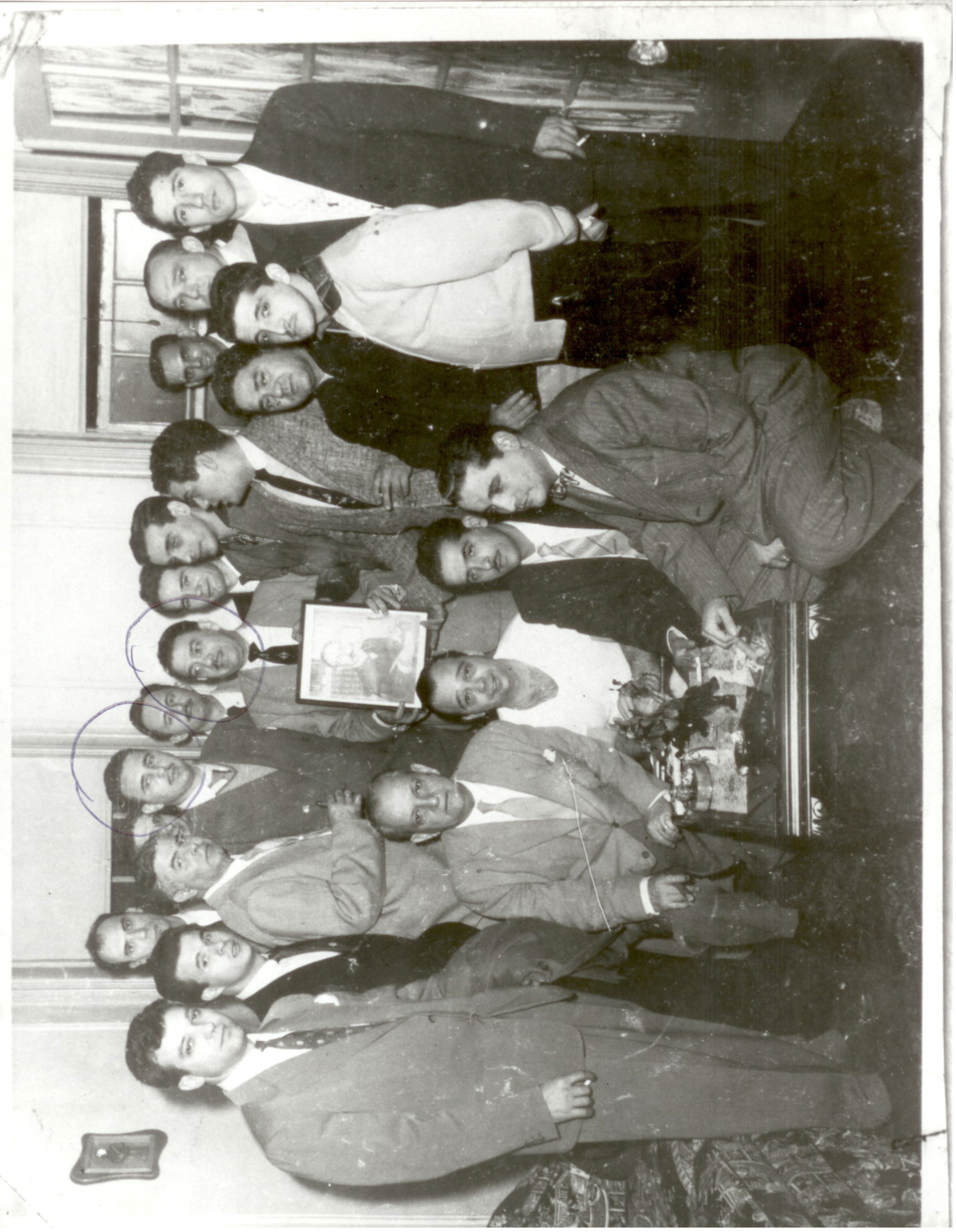 Vintage group portrait of about twenty men posed indoors, one holding a small framed photograph, two faces circled in ink
