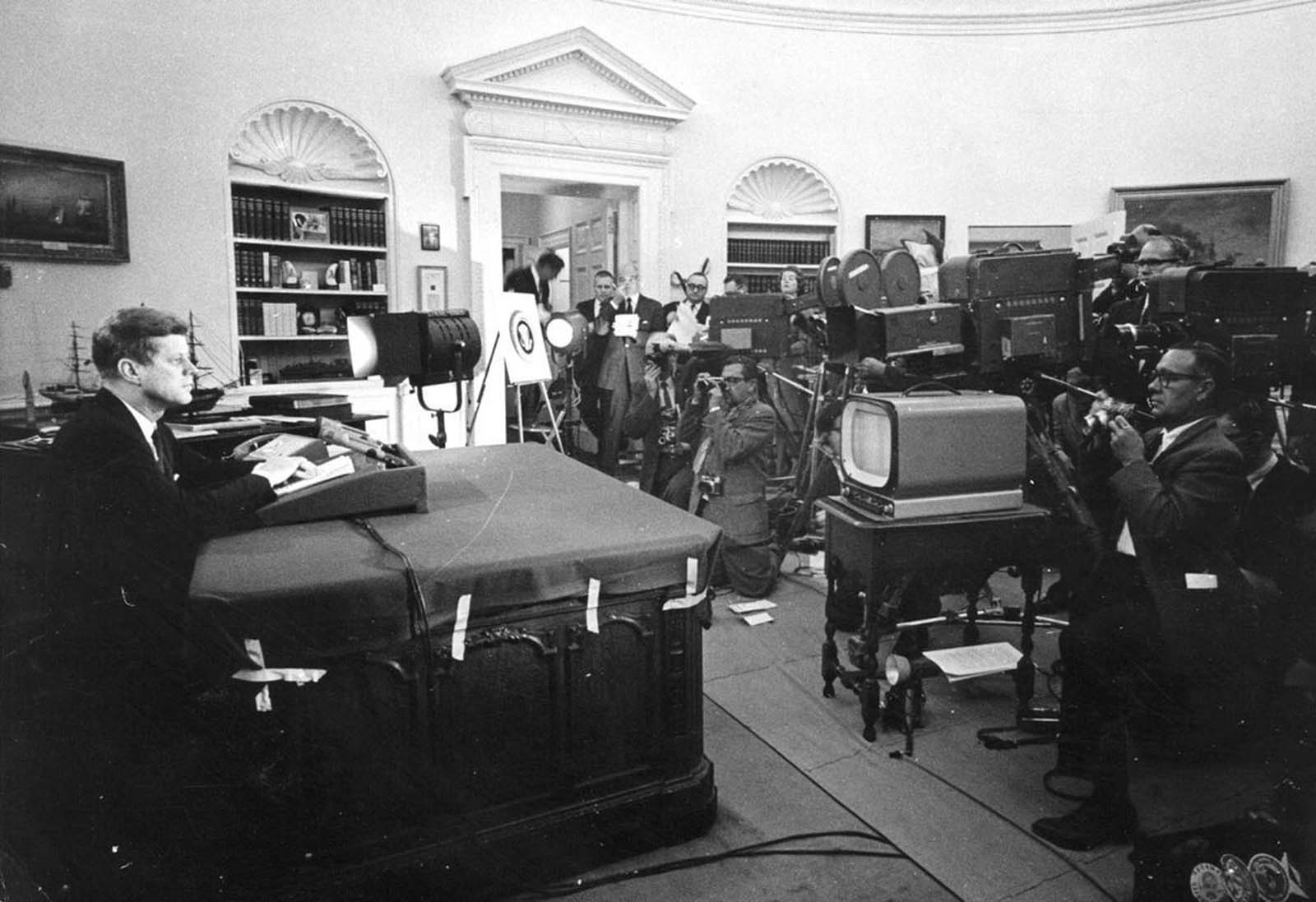 President Kennedy at his desk in the Oval Office facing television cameras and photographers