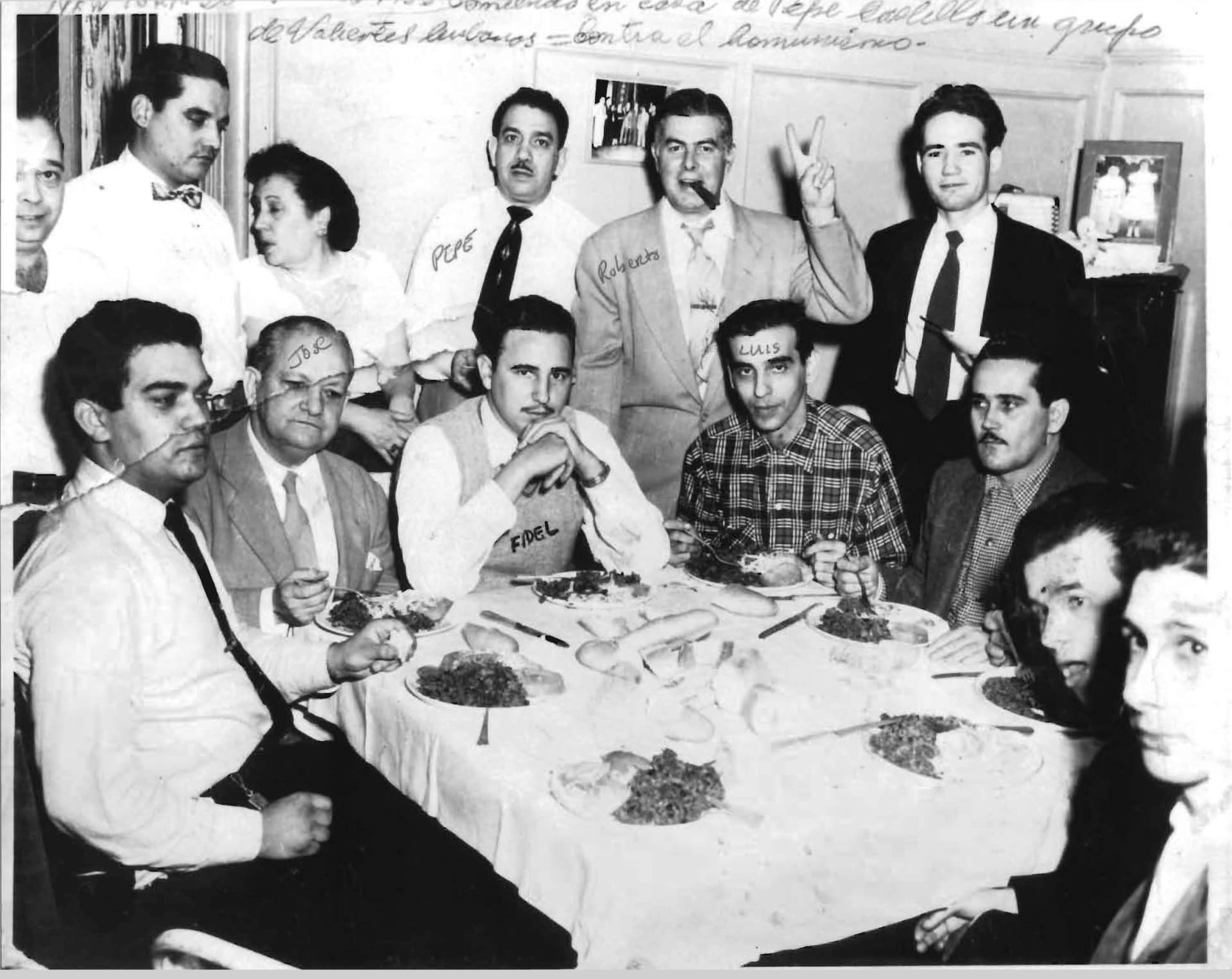Black-and-white photograph of men gathered around a dinner table with a handwritten Spanish inscription along the top margin