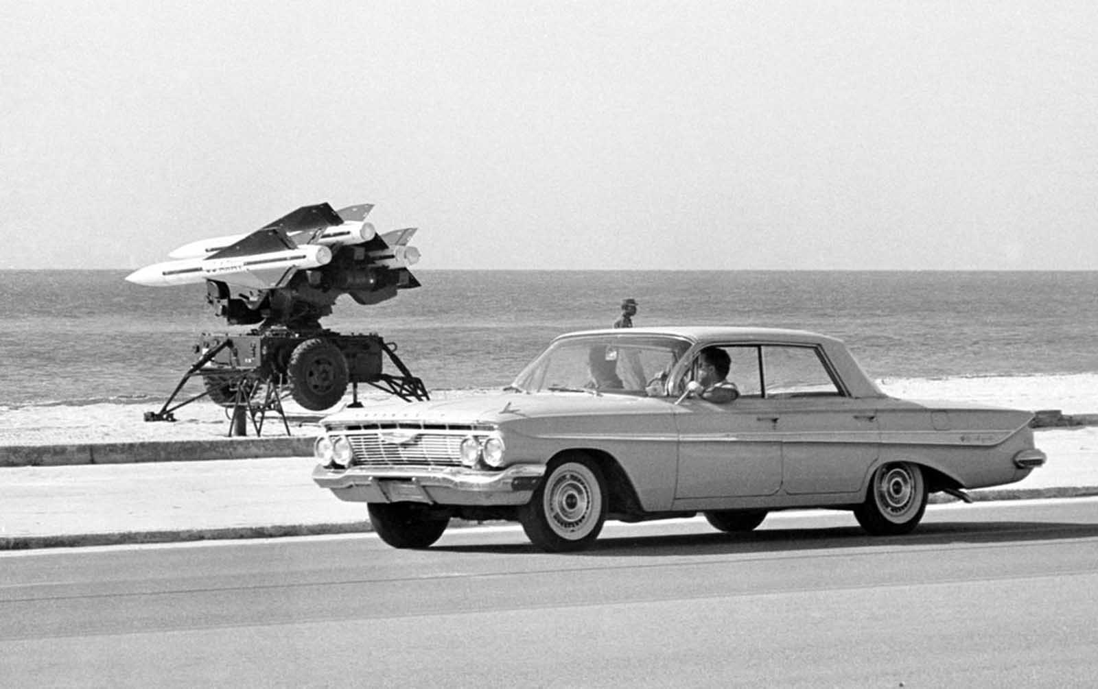 Black-and-white photograph of a Chevrolet sedan on a coastal road with a missile launcher carrying three missiles on the beach behind it