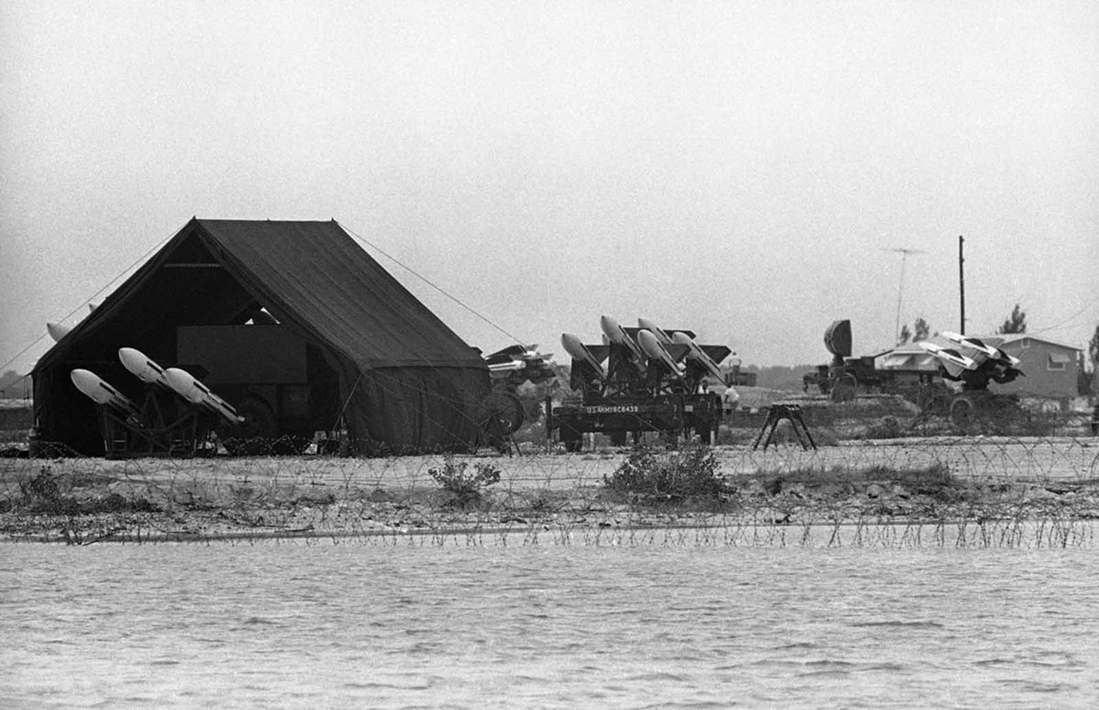 Black-and-white photograph of a coastal missile battery with loaded launchers, a tent and barbed wire in front of the water